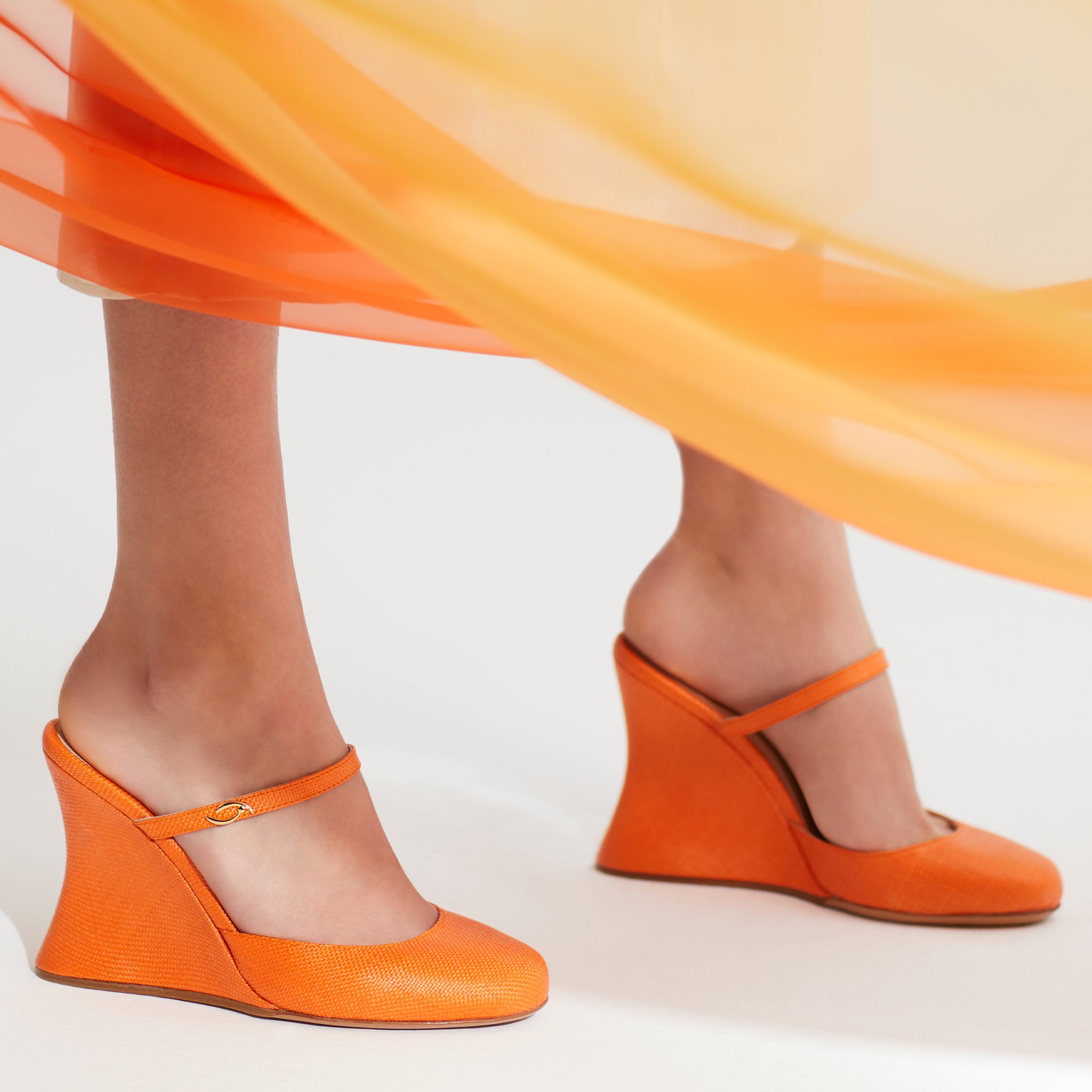 A pair of vibrant orange raffia Mary Jane mules with a signature hourglass wedge shape and gold jewel logo brooch. Raffia evening wedge for golden-hour dinners.
